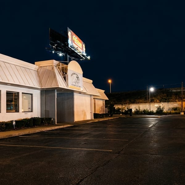 Exterior of a restaurant at night with a large parking lot and a billboard above.