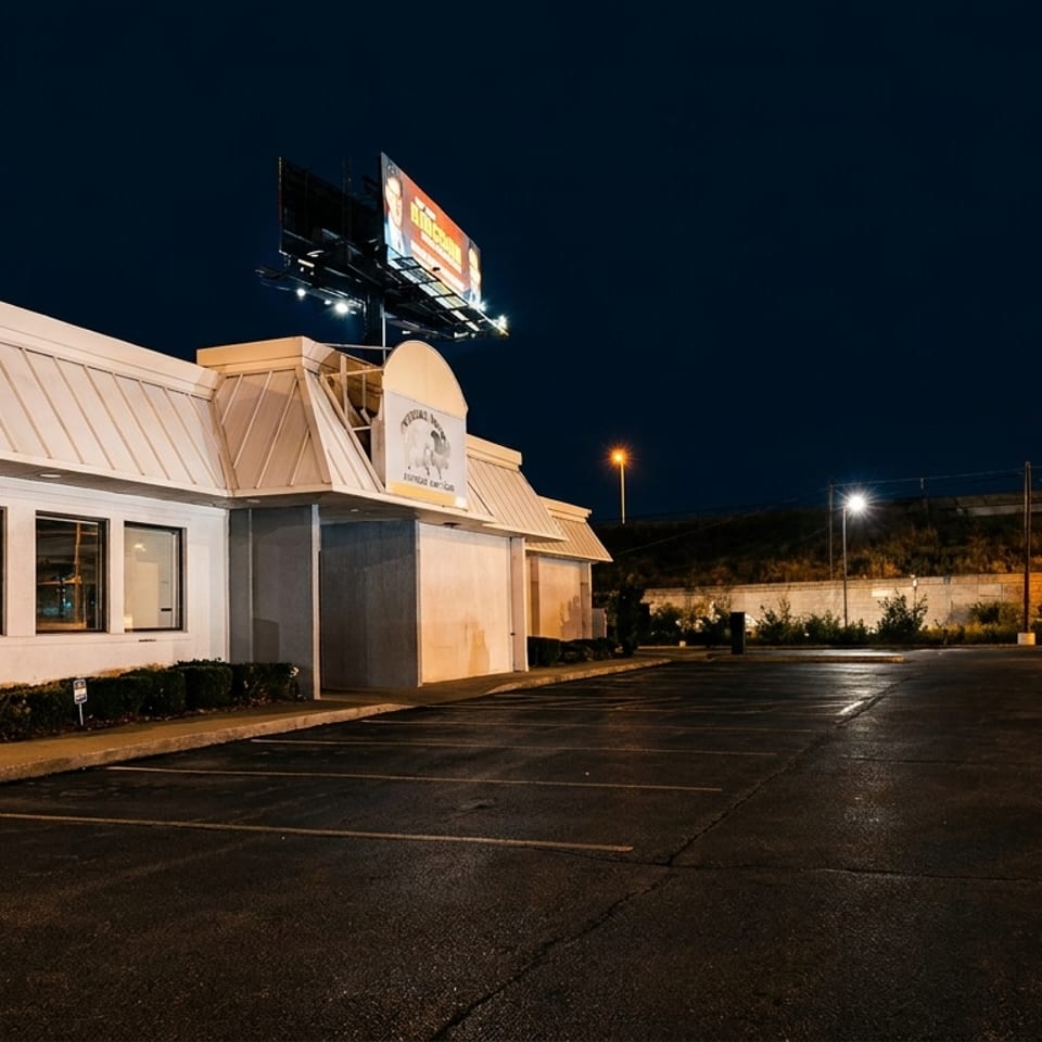 Exterior of a restaurant at night with a large parking lot and a billboard above.